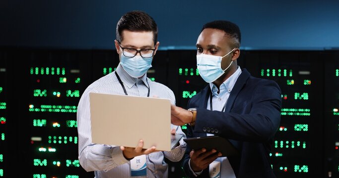 Caucasian and African American men with laptop and tablet device working in server room. Software engineers in data storage checking big data processors Male analytics co-workers working in datacenter