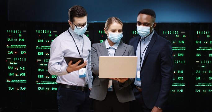 Caucasian and African American male and female analytics with laptop and tablet working in data storage. Software engineers in datacenter talking and checking big data processors. Colleagues concept