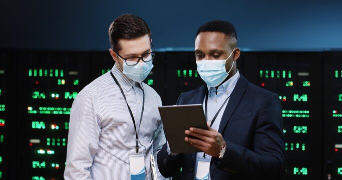Close up portrait of mixed-races male security analytics in medical masks typing on tablet and speaking standing in datacenter. Coworkers discussing technical issue in server room. Big data protection