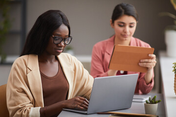 Portrait of contemporary African-American businesswoman using laptop at desk while managing successful company with female partner, copy space