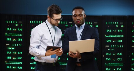 Caucasian and African American male analytics coworkers tapping on laptop and tablet and discussing data security issue working in data center. Man engineers speaking in data storage with server racks