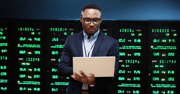 Portrait of handsome African American male security engineer standing in datacenter checking security of processors with digital information working in data storage and typing on laptop. Tech concept