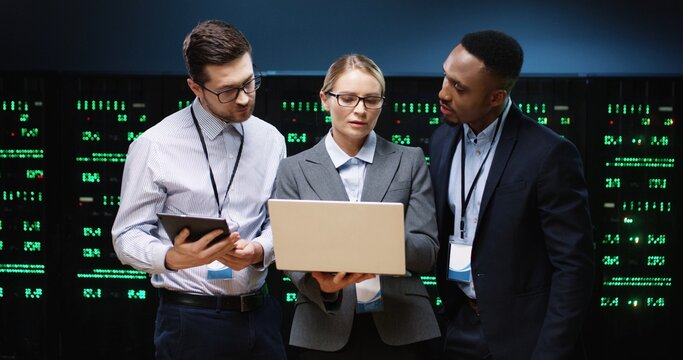 Portrait of mixed-races male and female analytics with laptop and tablet working in data center on cyber security issue. Engineers coworkers in server room talking and checking big data processors