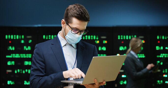 Caucasian man in mask with laptop working typing on computer in server room. Software male engineer in data storage check big data processors. Female analytic working on tablet on background. Close up