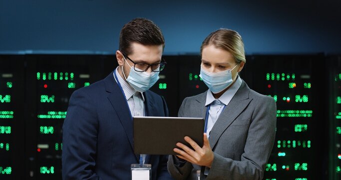 Close up portrait of Caucasian woman system administrator in mask standing in datacenter typing on tablet and speaking with male security engineer about data protection and digital information