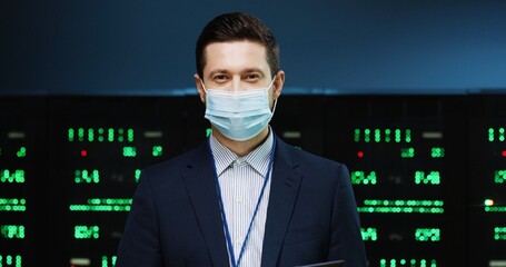 Close up portrait of Caucasian man system administrator in mask standing in High Tech big data center checking security of processors working with digital information and typing on tablet Work concept