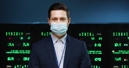 Close up portrait of Caucasian handsome male professional security engineer in medical mask stan ding in datacenter and looking at camera. Young happy man working in data storage. Server room concept