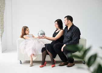 Young family of three sitting on white sofa in light room with green plant. Small daughter is giving christmas ornament to her mother. Stay home for winter holidays. Family values.