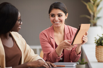 Portrait of smiling Middle-Eastern businesswoman looking at female colleague and pointing at tablet...