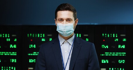 Close up portrait of Caucasian handsome male professional security engineer in medical mask stan ding in datacenter and looking at camera. Young happy man working in data storage. Server room concept