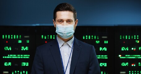 Close up portrait of Caucasian handsome male professional security engineer in medical mask stan ding in datacenter and looking at camera. Young happy man working in data storage. Server room concept