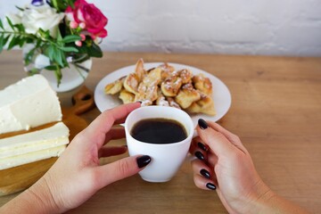 Cup of coffee in female hands, close-up. Homemade baked goods during quarantine. Homemade feta cheese on a wooden tabletop. Concept on the background of a white brick wall and a bouquet of roses.