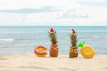 Family of funny attractive pineapples in stylish sunglasses on the sand against turquoise sea. Wearing christmas hats. Christmas and new year vacation concept on tropical beach. Family holiday. Bright