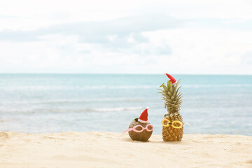  funny attractive pineapples and coconut in stylish sunglasses on the sand against turquoise sea. Wearing christmas hats. 