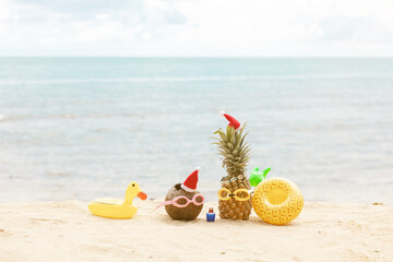  funny attractive pineapples and coconut in stylish sunglasses on the sand against turquoise sea. Wearing christmas hats. 