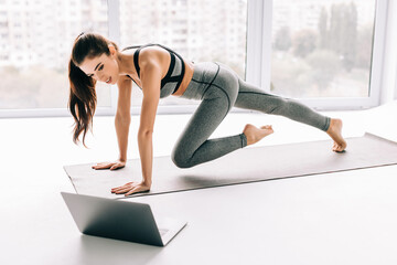 Strong sporty woman is doing working out at home and doing plank in front of her laptop, wearing sport outfit