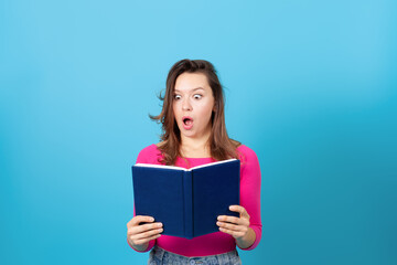 a close up of surprised young woman with mouth open holding a diary or a book , isolated on blue background