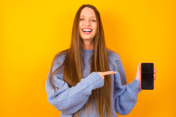 Attractive cheerful Young beautiful Caucasian woman wearing blue sweater against yellow wall holding in hands cell showing black screen