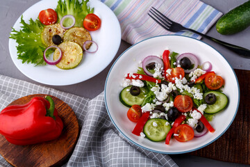 White plate with grilled zucchini with cherry tomatoes on lettuce and Greek salad on this table on a napkin with a fork near vegetables.