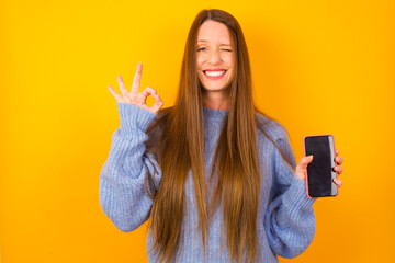Young beautiful Caucasian woman wearing blue sweater against yellow wall holding in hands cell...