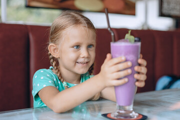 A little cute girl is looking at a large glass of milkshake and enjoying a sweet dessert in a local cozy cafe. Portrait.