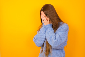 Sad Young beautiful Caucasian woman wearing blue sweater against yellow wall crying covering her face with her hands.