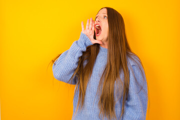 Young beautiful Caucasian woman wearing blue sweater against yellow wall shouting and screaming loud to side with hand on mouth. Communication concept.