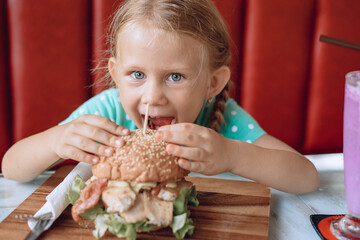 A little pretty cute girl with blond hair and blue eyes is trying to bite a big burger in a local cafe. Portrait.