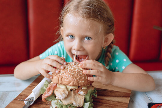 A Little Pretty Cute Girl With Blond Hair And Blue Eyes Is Trying To Bite A Big Burger In A Local Cafe. Portrait.