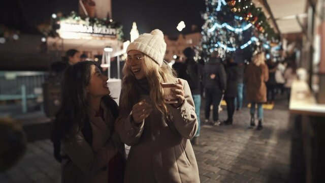 Pretty young girl surprising friend, running into her from behind. Teen friends standing in a middle of christmas fare, drinking coffee, having great time together.