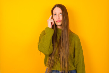 Young beautiful Caucasian woman wearing green sweater against yellow wall tries to memorize something, keeps fore finger on temple, reminds information for exam,