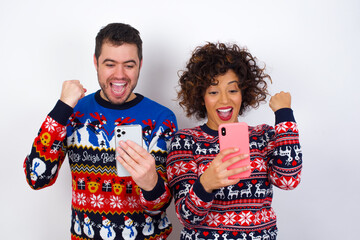 charming cheerful Young couple wearing Christmas sweater standing against white wall holding in hands cell reading browsing