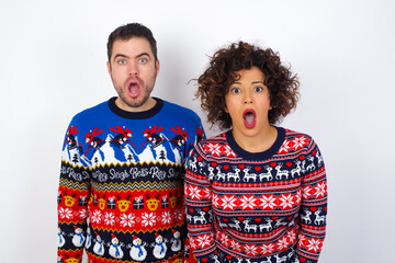 Oh my God. Surprised Young couple wearing Christmas sweater standing against white wall stares at camera with shocked expression exclaims with unexpectedness,