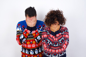 Young couple wearing Christmas sweater standing against white wall covering her face with her hands, being devastated and crying. Sad concept