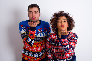 Young couple wearing Christmas sweater standing against white wall looking at the camera blowing a kiss with hand on air being lovely and sexy. Love expression.
