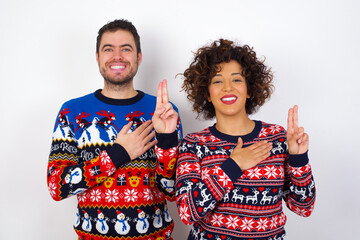 Young couple wearing Christmas sweater standing against white wall smiling swearing with hand on chest and fingers up, making a loyalty promise oath.