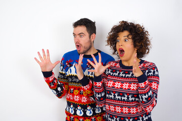 Young couple wearing Christmas sweater standing against white wall shouts loud, keeps eyes opened and hands tense.