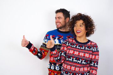 Young couple wearing Christmas sweater standing against white wall Looking proud, smiling doing thumbs up gesture to the side. Good job!