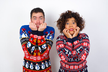 Fearful Young couple wearing Christmas sweater standing against white wall keeps hands near mouth, feels frightened and scared,  has a phobia,  Shock and frighted concept.