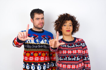 Young couple wearing Christmas sweater standing against white wall making fun of people with fingers on forehead doing loser gesture mocking and insulting.