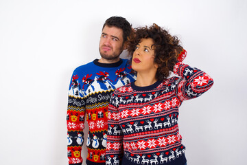 Young couple wearing Christmas sweater standing against white wall confuse and wonder about question. Uncertain with doubt, thinking with hand on head. Pensive concept.