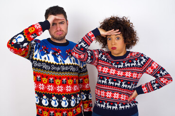 Young couple wearing Christmas sweater standing against white wall having problems, worried and stressed holds hand on forehead.