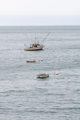 A fishing trawler is at anchor in a ocean bay. It has to poles out on each side of the boat. A buoy...
