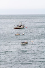 A fishing trawler is at anchor in a ocean bay. It has to poles out on each side of the boat. A buoy is between the boat and a small dingy. The water is grey.