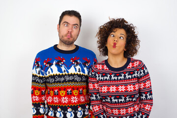 Young couple wearing Christmas sweater standing against white wall making fish face with lips, crazy and comical gesture. Funny expression.
