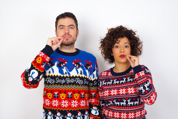 Young couple wearing Christmas sweater standing against white wall mouth and lips shut as zip with fingers. Secret and silent, taboo talking.