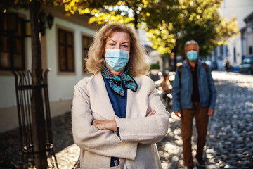 Senior woman with a protective mask on standing with arms crossed in an old part of the city. In background is senior man walking, having protective mask. We have to be responsible for our old ones.