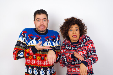 Young couple wearing Christmas sweater standing against white wall In hurry pointing to watch time,...