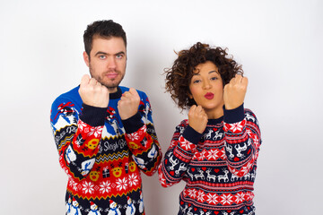 Young couple wearing Christmas sweater standing against white wall Ready to fight with fist defense gesture, angry and upset face, afraid of problem.