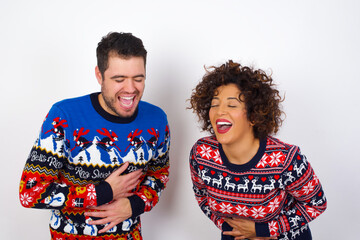 Young couple wearing Christmas sweater standing against white wall smiling and laughing hard out loud because funny crazy joke with hands on body.